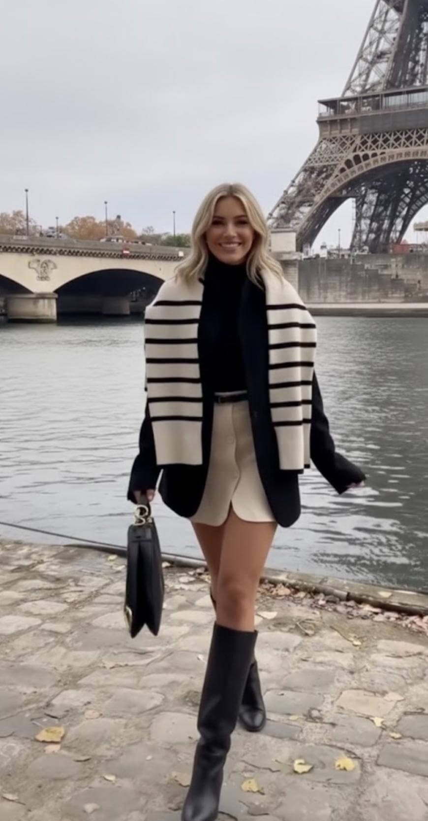 A woman wearing a black turtleneck, cream skirt, striped scarf, and knee-high boots walks by the Seine River with the Eiffel Tower in the background on an overcast day in Paris.
