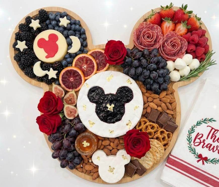 A Mickey Mouse-shaped charcuterie board with cheese, berries, grapes, figs, nuts, cured meats, crackers, pretzels, and fresh red roses. A napkin with The Brave is on the side.