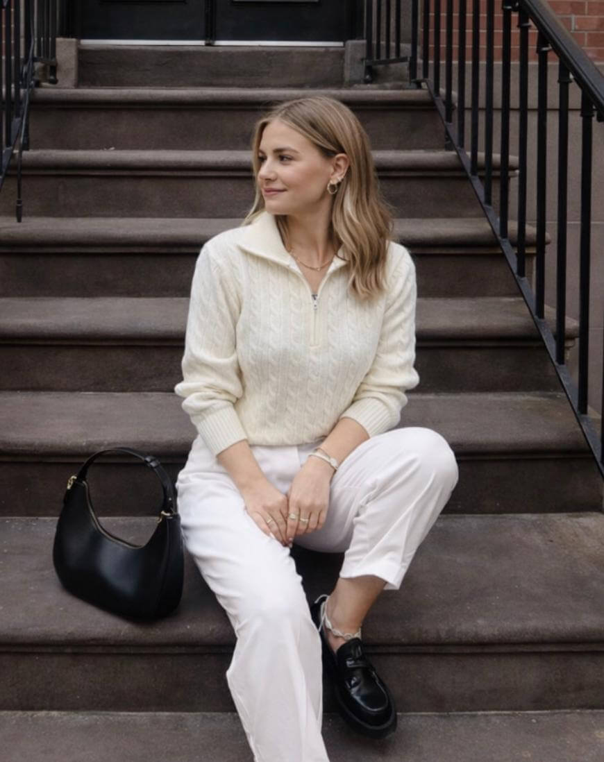 A woman with light brown hair sits on outdoor steps, wearing a cream cable-knit sweater, white pants, and black shoes. She has a black handbag beside her and looks to the side, smiling slightly.