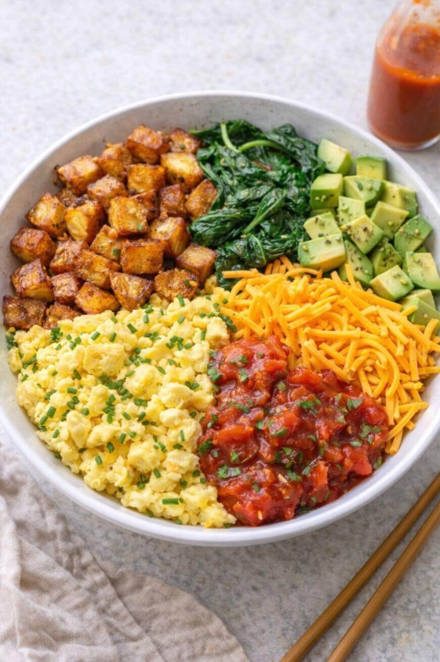 A bowl filled with scrambled eggs, roasted potatoes, sautéed spinach, diced avocado, shredded cheddar cheese, and salsa, arranged in colorful sections. A bottle of hot sauce is visible in the background.