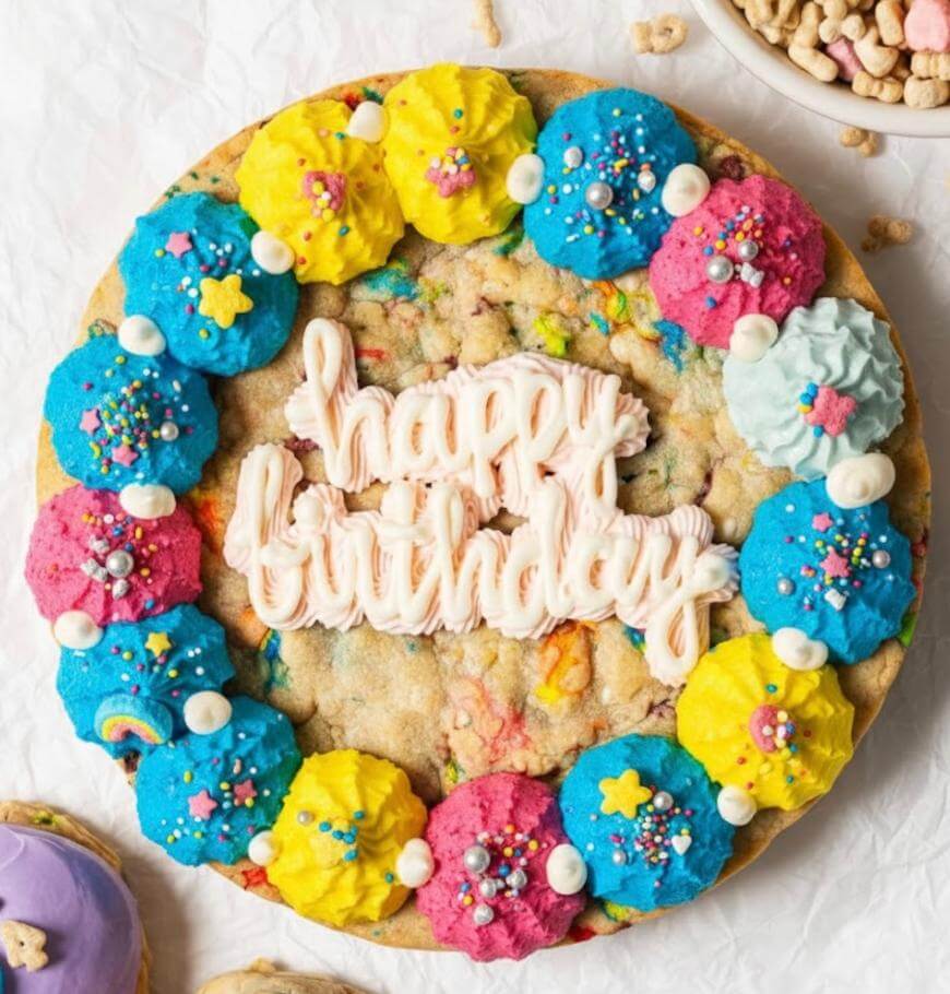 A large cookie cake decorated with colorful blue, pink, and yellow frosting dollops, sprinkles, and white stars. In the center, white frosting spells out happy birthday.