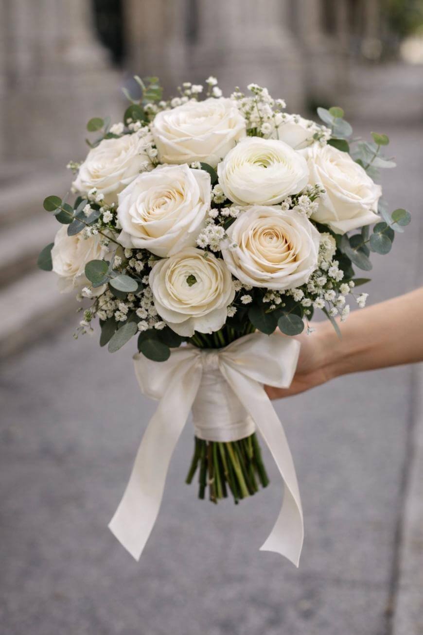 A hand holds a bouquet of white roses, ranunculus, and babys breath, accented with eucalyptus leaves and tied with a white ribbon. The background is softly blurred and appears to be outdoors.