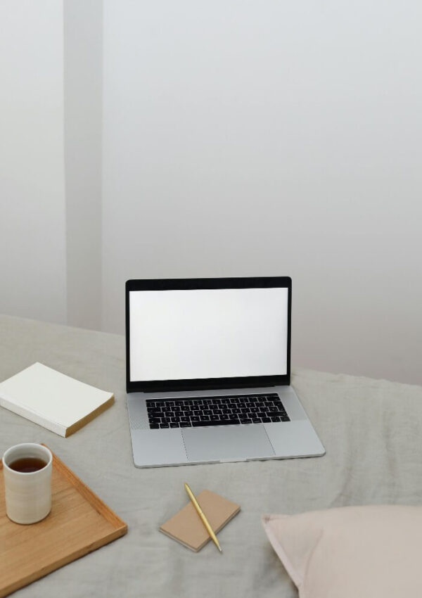 A laptop with a blank screen sits on a bed next to a notebook, a pencil, a tray holding a cup of coffee, and two small notepads. The setting is minimal and cozy with soft lighting.