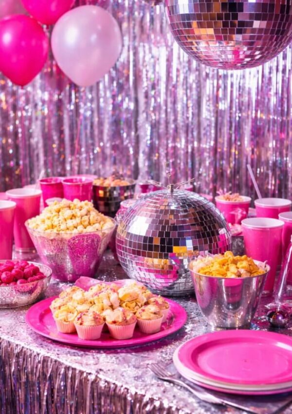 A party table decorated with shiny silver tinsel, disco balls, and pink tableware. Snacks like popcorn and cupcakes are served, with pink balloons and drinks in the background, creating a festive, vibrant atmosphere.