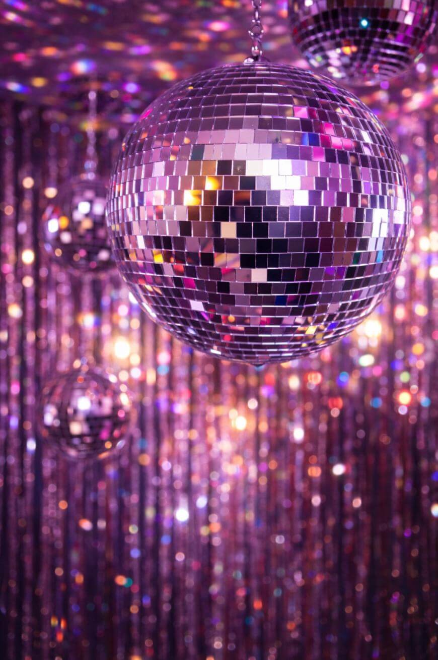 A close-up of a shiny disco ball reflecting colorful lights, with a sparkling, purple-toned tinsel backdrop and more disco balls in the background.