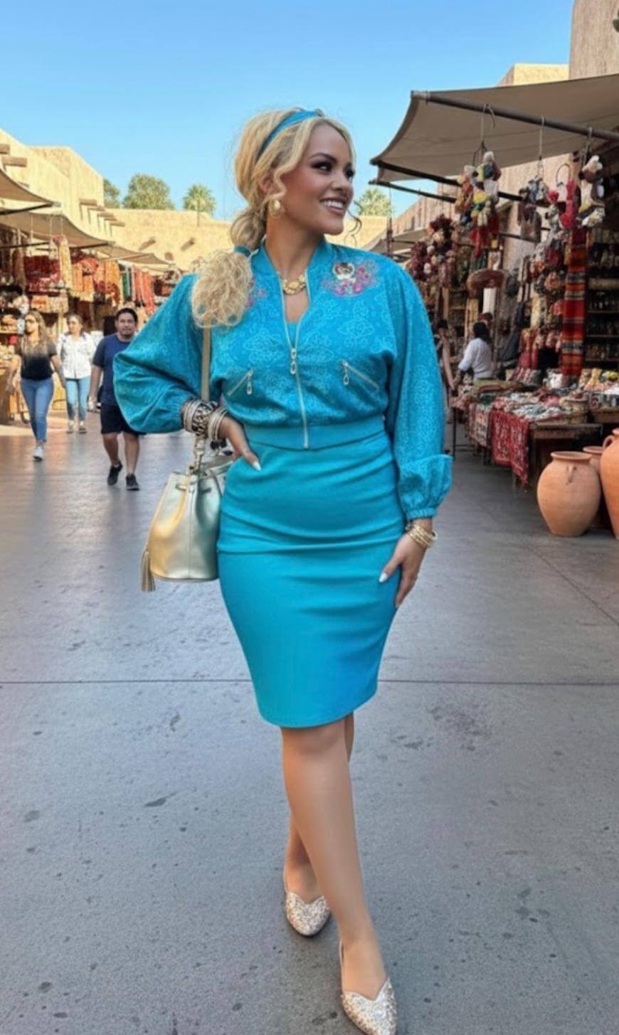 A woman in a blue blouse and matching skirt walks through an outdoor market, smiling. She carries a silver handbag and wears jewelry, with her blond hair styled in a ponytail and headband. Stalls with pottery and textiles line the background.