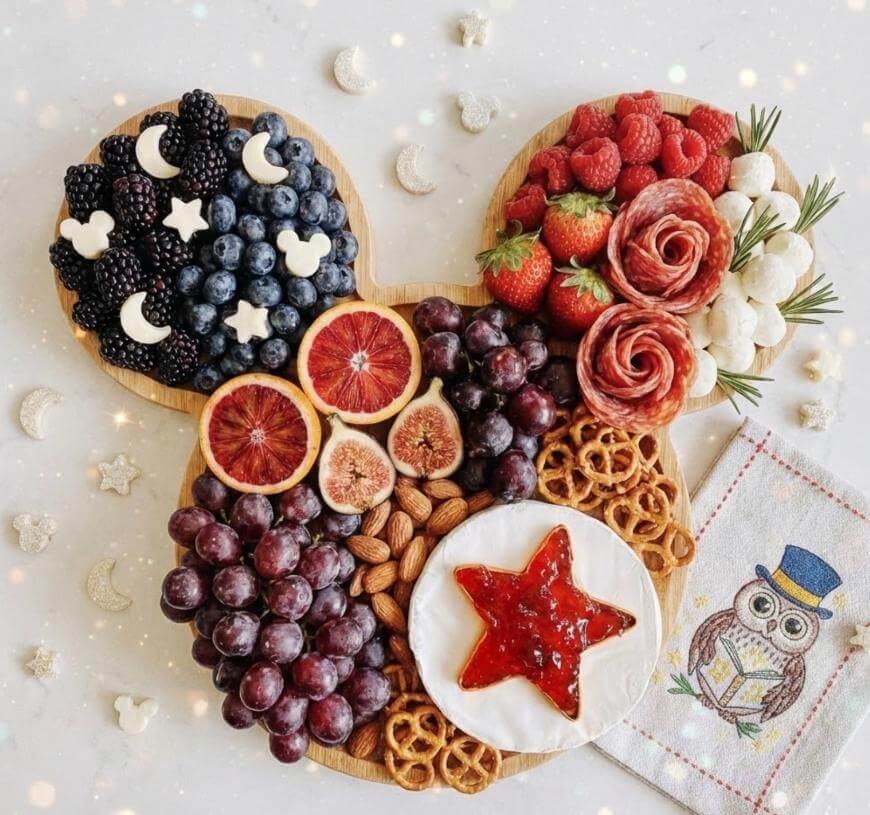 A charcuterie board shaped like Mickey Mouse is filled with berries, grapes, figs, almonds, pretzels, mozzarella balls, salami roses, star-shaped jam on brie, and Mickey-shaped cheese, with a decorative owl napkin nearby.