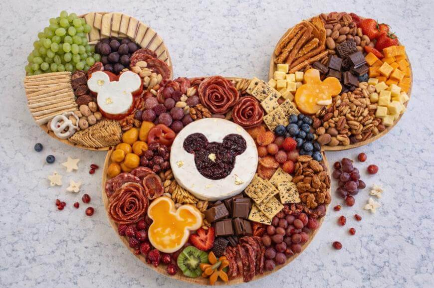 A large charcuterie board shaped like Mickey Mouse features assorted cheeses, crackers, fruits, nuts, and meats, with cheese and jam cut-outs in the shape of Mickey Mouse heads on each section.
