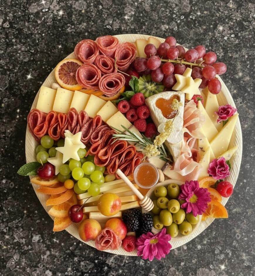 A round platter with assorted cheeses, salami, green and red grapes, apple slices, olives, raspberries, blackberries, dried apricots, honey, crackers, a wedge of brie with jam, and edible flowers on a dark countertop.