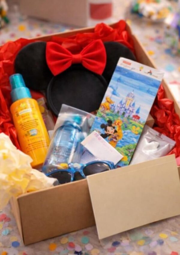 A gift box contains a pair of Minnie Mouse ears with a red bow, sunscreen, a water bottle, sunglasses, Disney-themed tickets, and other small items, all nestled in red tissue paper with confetti around.