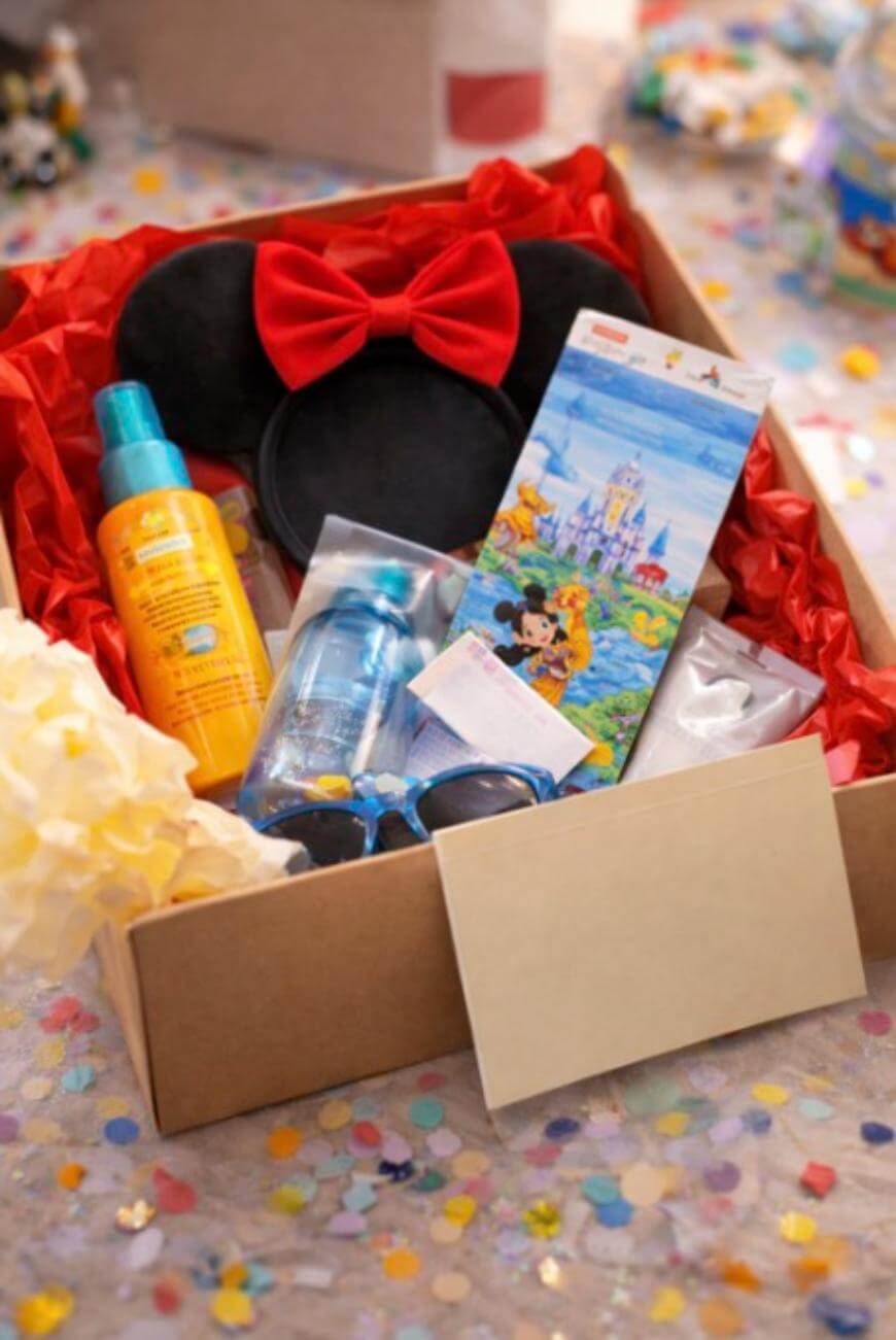 A gift box filled with Minnie Mouse ear headband, sunscreen, bottled water, sunglasses, a map with Disney characters, and an envelope, surrounded by confetti on a table.