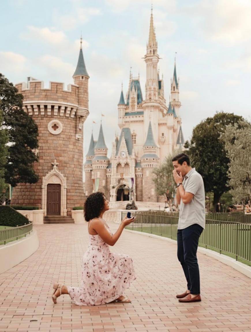 A woman kneels and proposes to a surprised man in front of a fairytale castle at a theme park, surrounded by greenery and towers, under a partly cloudy sky.