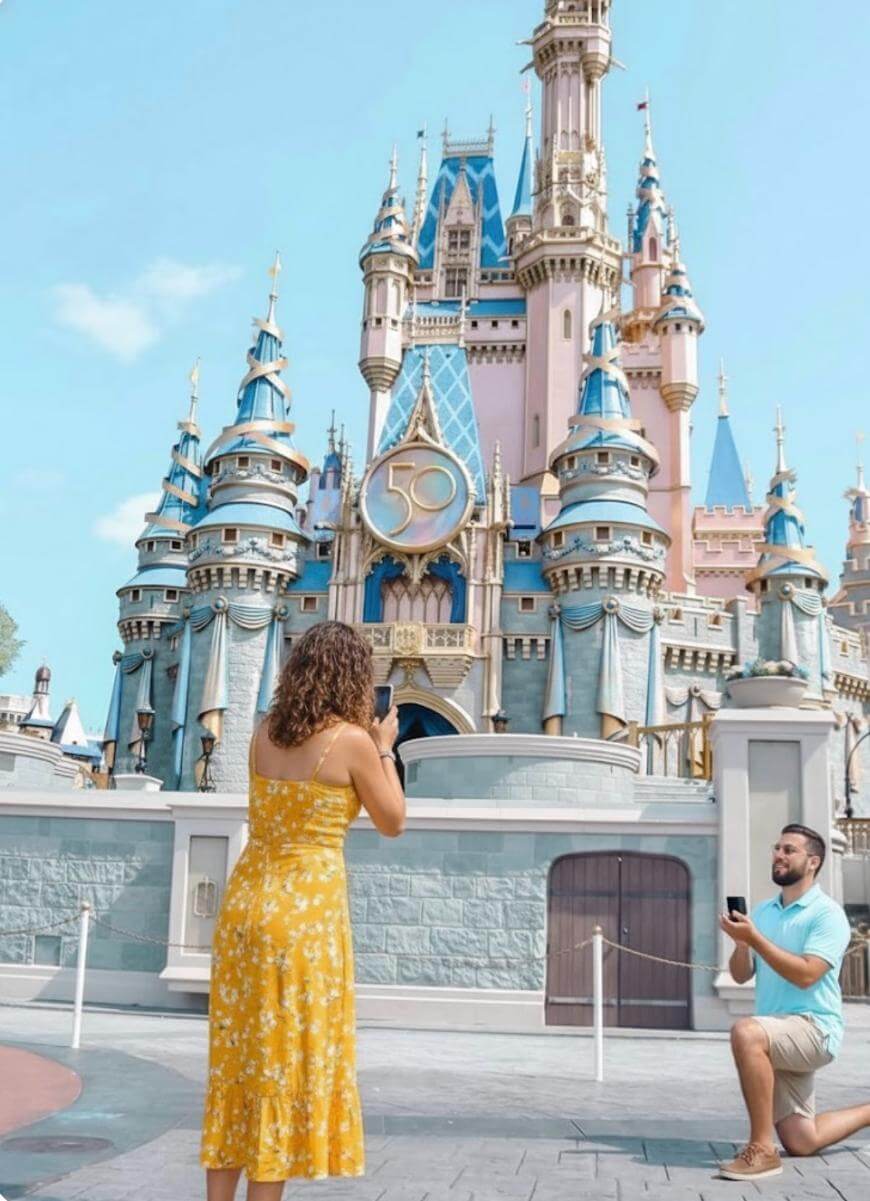A man kneels and proposes to a woman in a yellow dress in front of Cinderella’s Castle at Disney World, decorated with a gold 50 for the park’s anniversary.