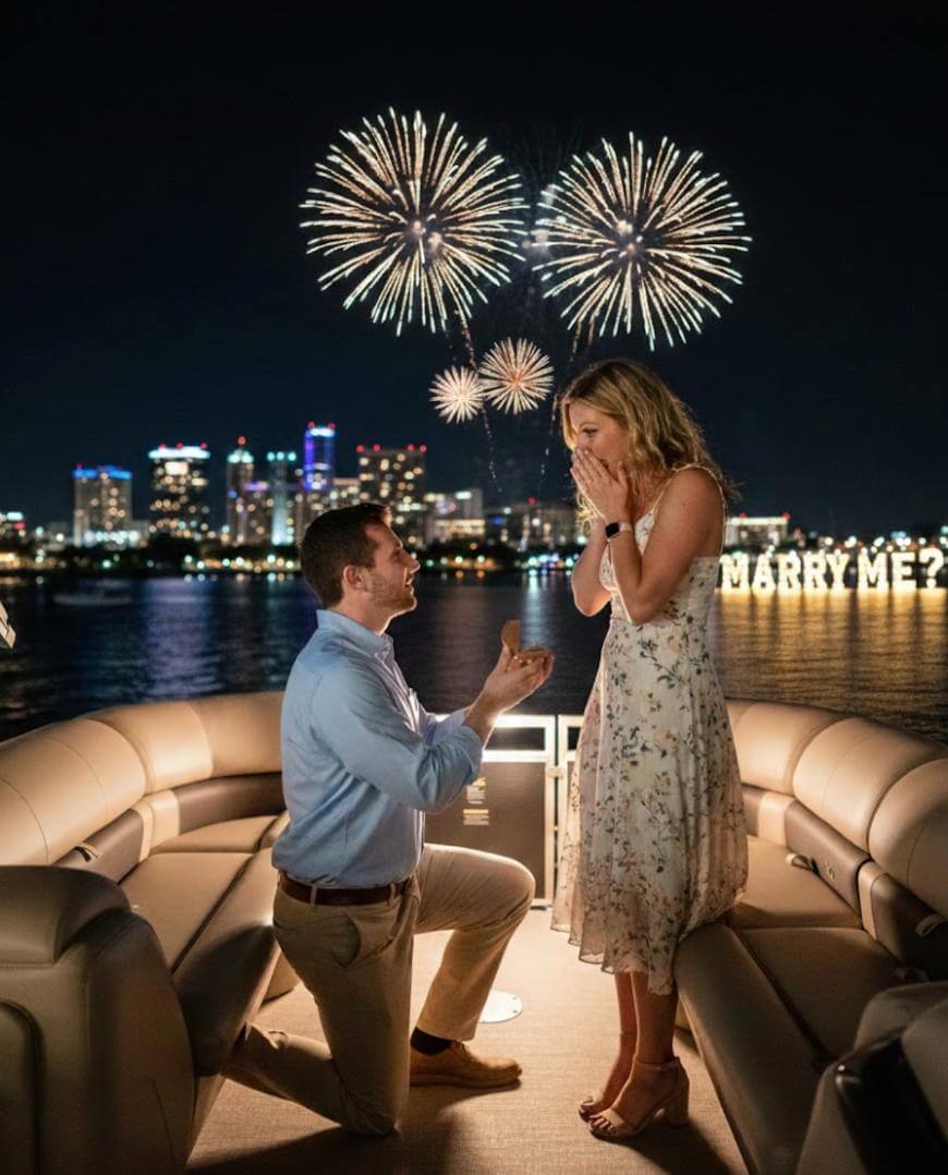 A man kneels on a boat at night, holding an open ring box, proposing to a surprised woman. Fireworks light up the sky, and illuminated MARRY ME? letters appear in the background with a city skyline.