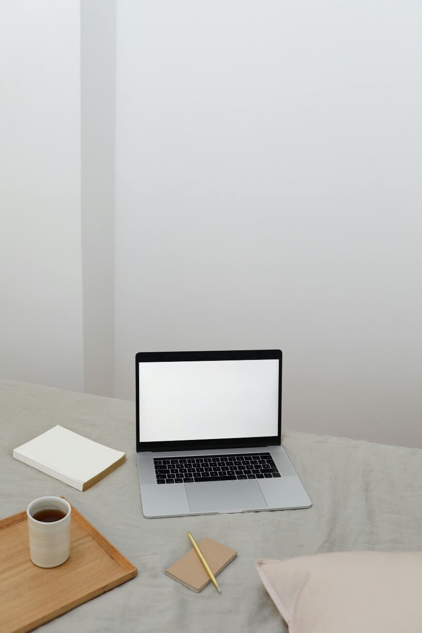 A laptop with a blank white screen sits on a bed beside a closed book, a notepad with a pencil, and a wooden tray holding a cup of coffee. The setting appears minimalistic and cozy.