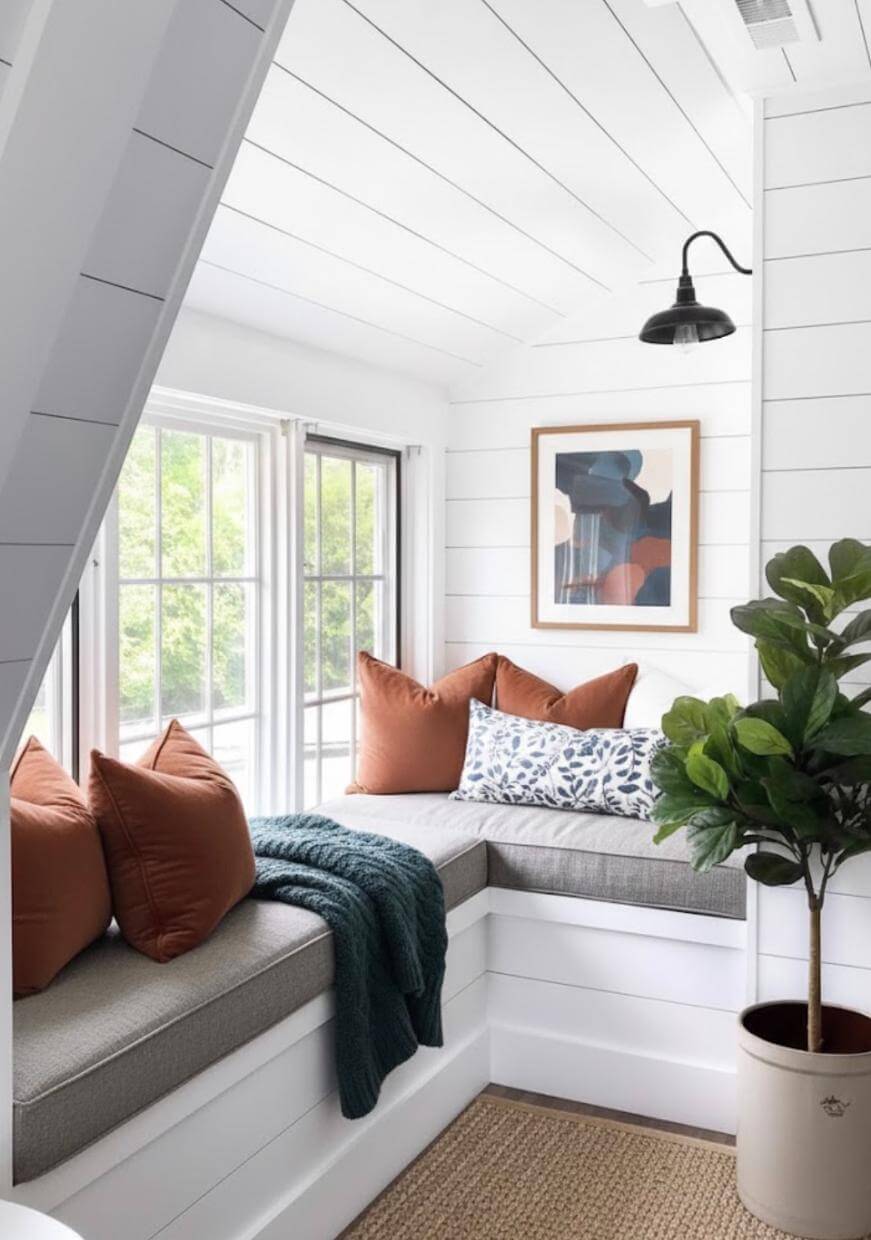 A cozy window nook with gray cushioned seating, rust-colored and patterned pillows, a green throw blanket, a leafy potted plant, a framed abstract artwork, and a black wall sconce against white shiplap walls.