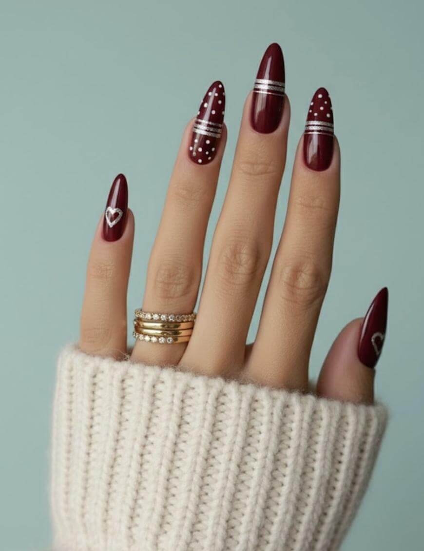 A hand with long, almond-shaped maroon nails featuring white designs: stripes, dots, and hearts. The person wears gold rings and a cream knit sweater against a light blue background.