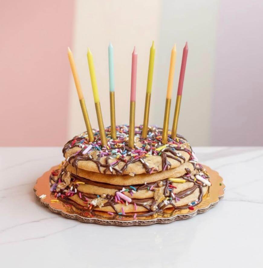 A stack of large cookies drizzled with chocolate and topped with colorful sprinkles serves as a cake. Seven pastel-colored candles are placed on top. The background features soft, pastel vertical stripes.