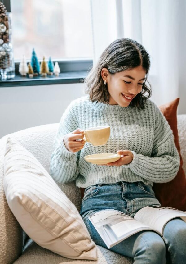 A young woman with shoulder-length hair, wearing a light blue sweater and jeans, sits on a cozy couch holding a cup and saucer, smiling while reading an open magazine. A window with decorations is in the background.