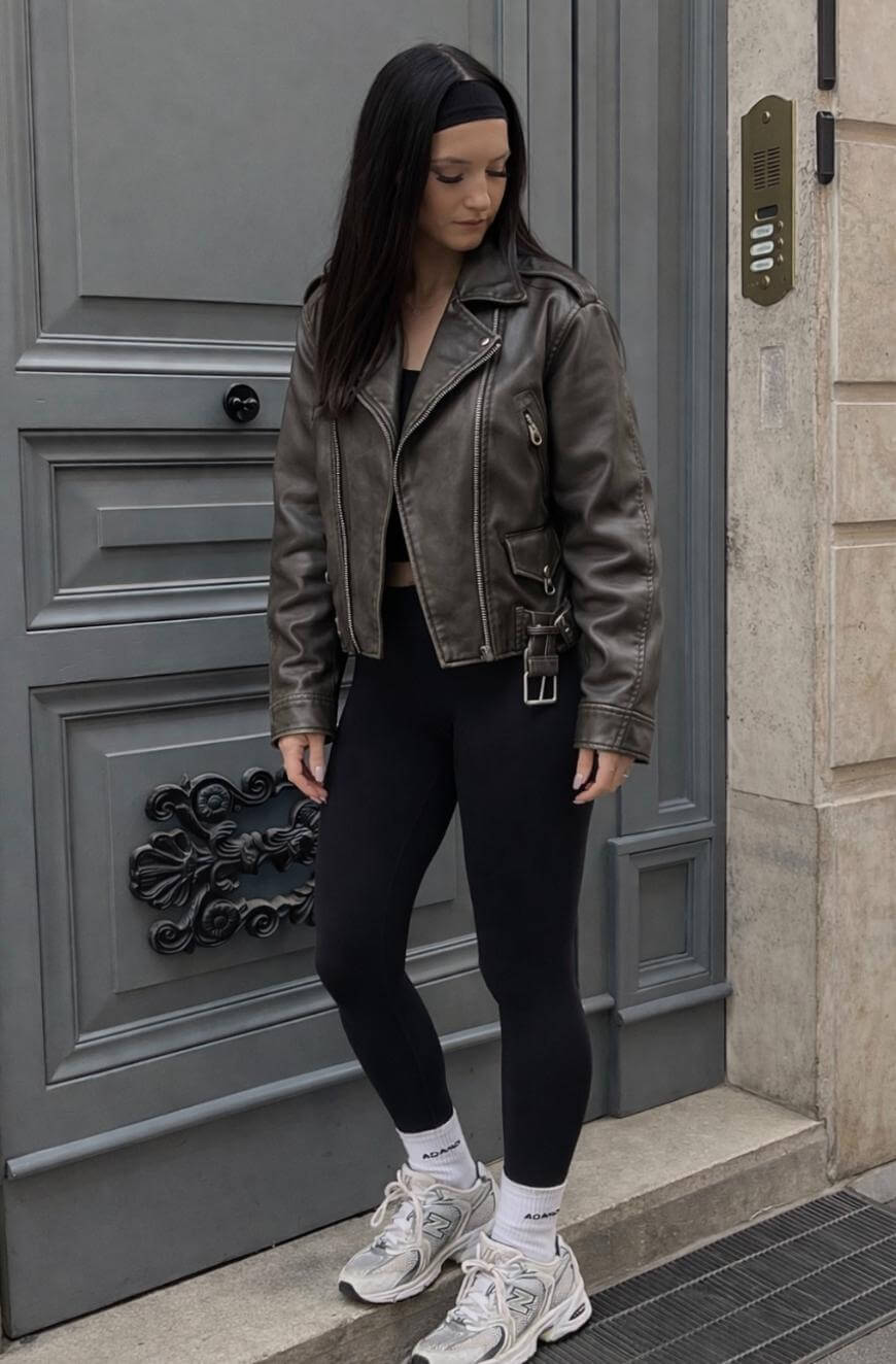 A woman with long dark hair wearing a black leather jacket, black leggings, white socks, and white sneakers stands on a city sidewalk in front of a large gray door, looking down to her left.