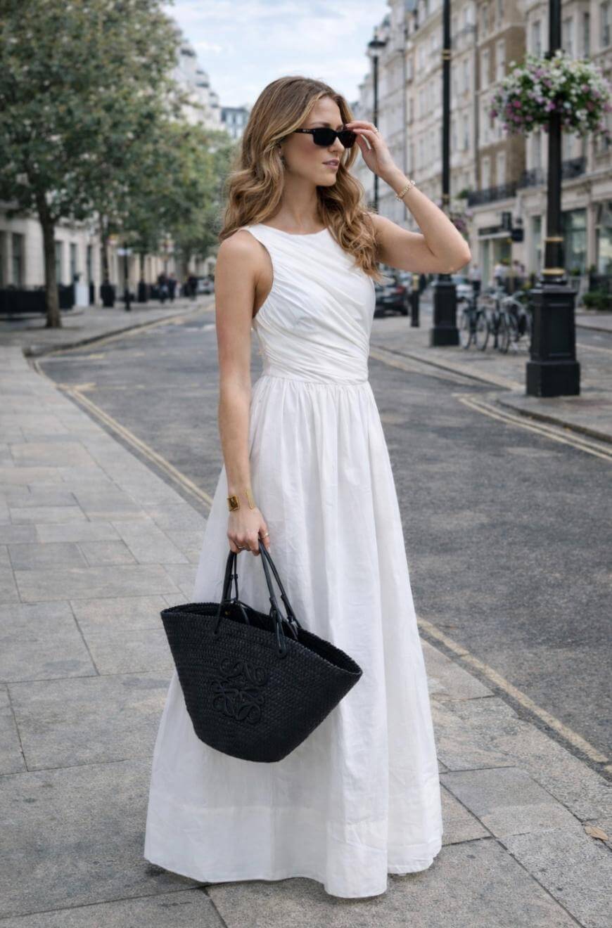 A woman in a sleeveless white maxi dress stands on a city street, wearing sunglasses and holding a black woven tote bag. Trees, bicycles, and buildings line the background.