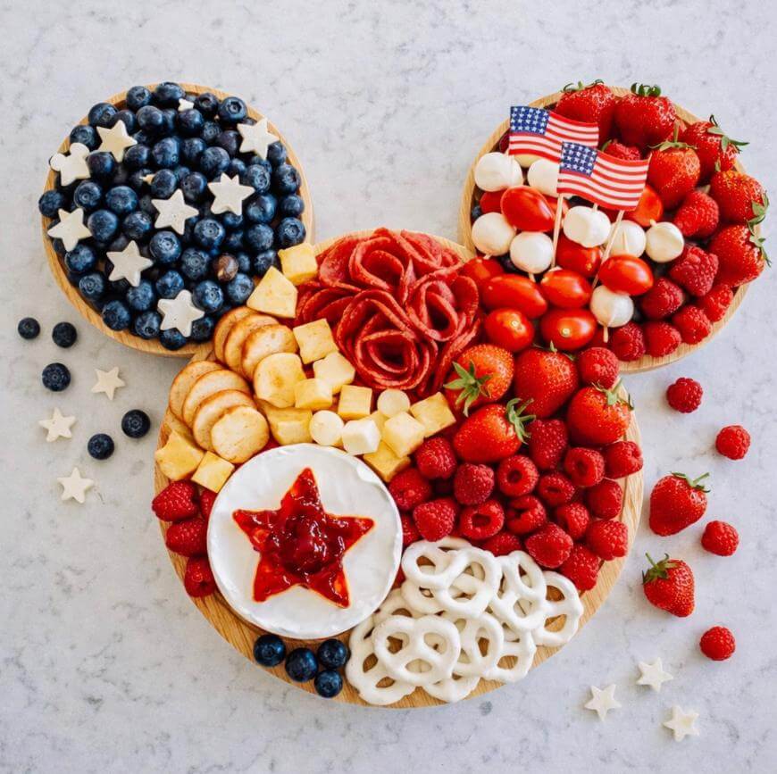 A snack tray arranged in the shape of Mickey Mouse, with sections of blueberries, cheese, strawberries, tomatoes, melon, meat, pretzels, and raspberries; American flags and star-shaped cheese decorate the platter.