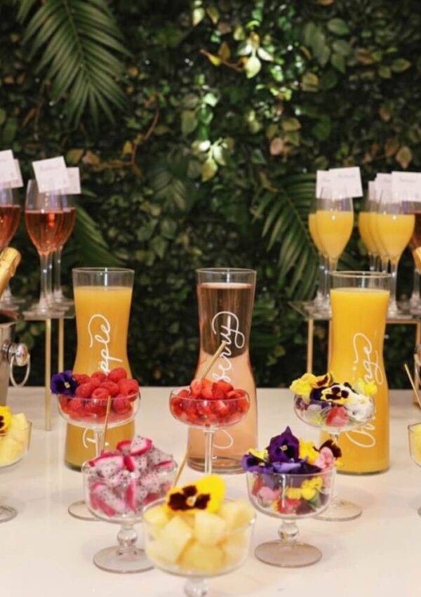 A colorful brunch setup featuring glasses of orange juice, champagne bottles in ice buckets, fruit bowls with edible flowers, and flutes of sparkling drinks against a lush green leafy background.