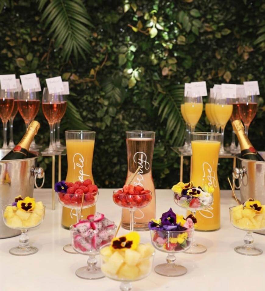A mimosa bar setup with champagne bottles, glasses of orange juice, rosé, and garnishes like fresh fruit and edible flowers, arranged on a white table against a leafy green backdrop.