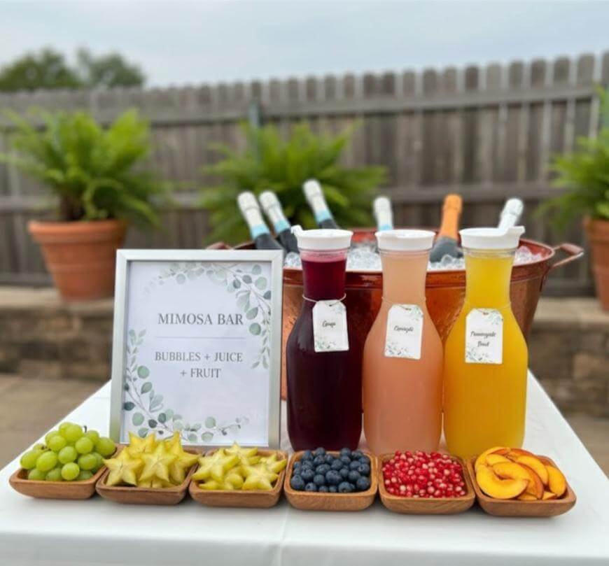 A mimosa bar setup on a white table features bottles of champagne in an ice bucket, three carafes of grape, orange, and pineapple juice, and bowls of grapes, starfruit, blueberries, pomegranate seeds, and peach slices.