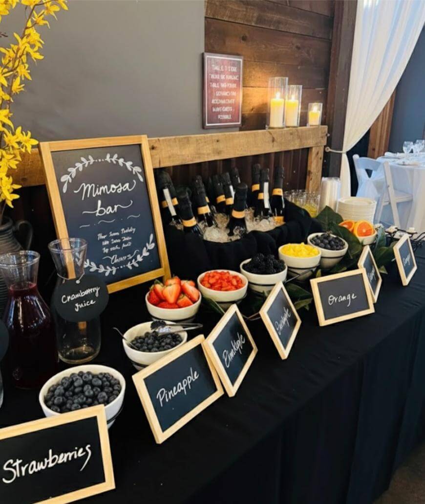 A mimosa bar setup with bottles in ice, carafes of juice, various bowls of fruit including strawberries, blueberries, pineapple, oranges, and blackberries, with chalkboard labels and candles in the background.