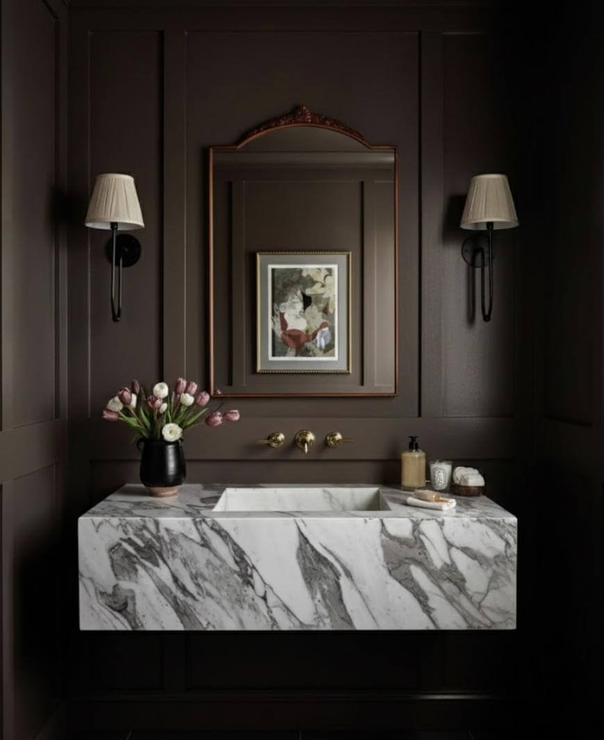 A dark brown bathroom features a marble sink, a vase of tulips, wall-mounted gold faucet, two sconces with beige shades, a mirror with wooden trim, and framed art above the sink. Soap and decor items are on the counter.