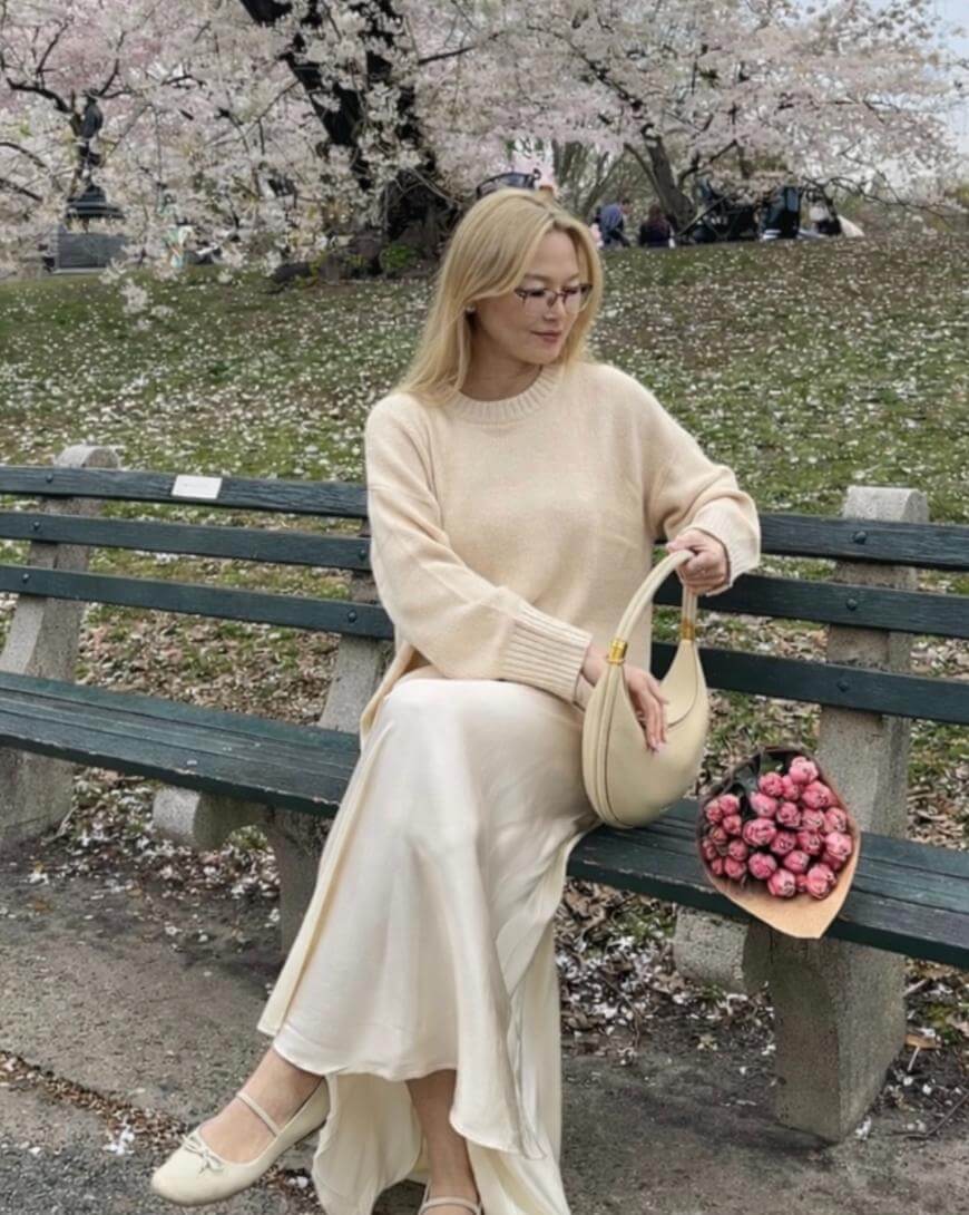 A woman in a cream sweater and long skirt sits on a park bench under blooming cherry blossom trees, holding a cream handbag. A bouquet of pink flowers rests beside her on the bench.