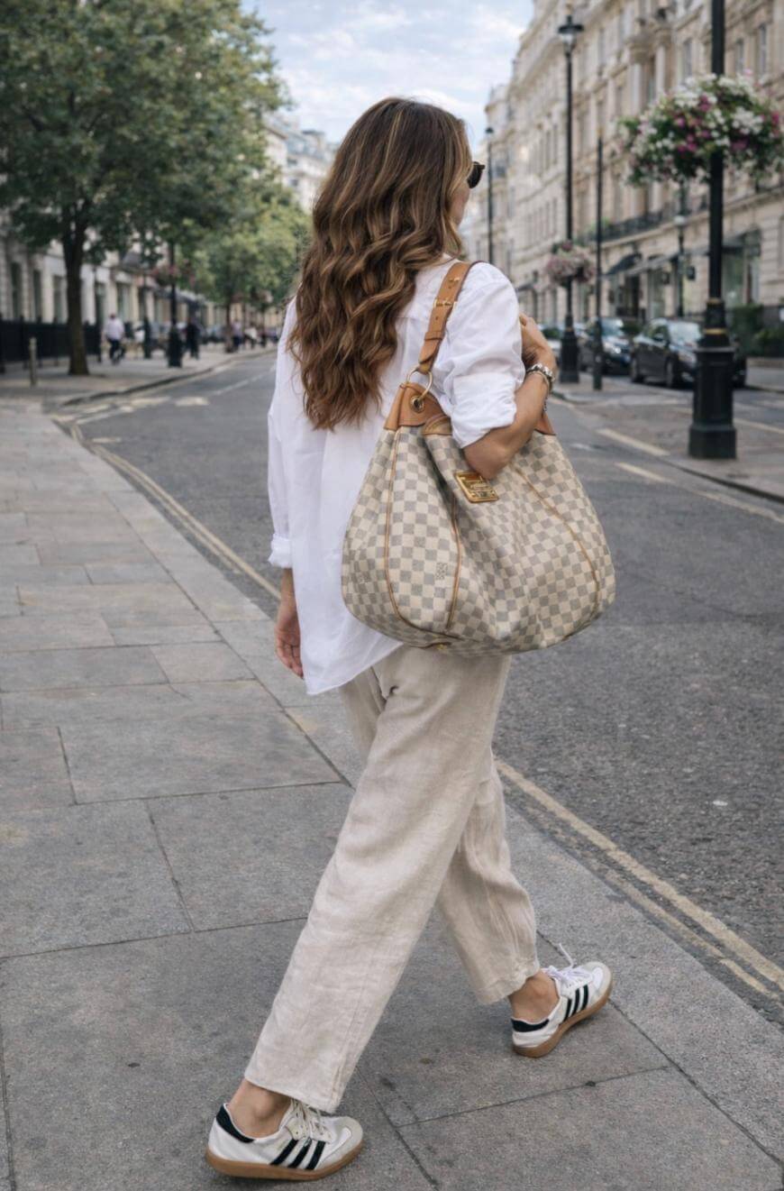 A woman with wavy brown hair, wearing a white shirt, beige pants, and black-and-white Adidas sneakers, walks on a city sidewalk carrying a large checkered shoulder bag. Trees and buildings line the street.