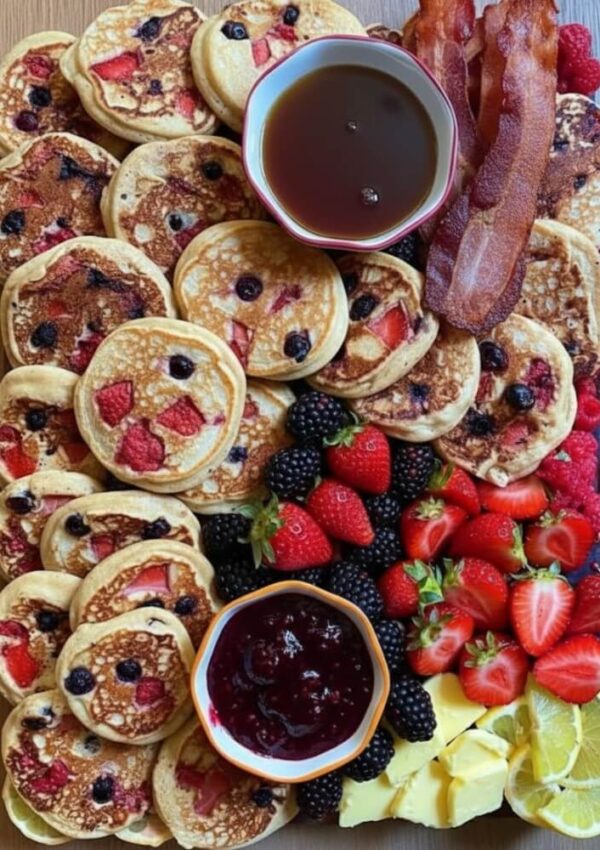 A wooden board topped with stacks of mini pancakes with berries, bowls of syrup and jam, crispy bacon, blackberries, strawberries, butter slices, and lemon wedges arranged neatly.