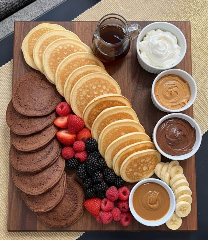 A wooden board with chocolate and plain pancakes, fresh berries, banana slices, and bowls of whipped cream, peanut butter, chocolate spread, and syrup arranged neatly for serving.