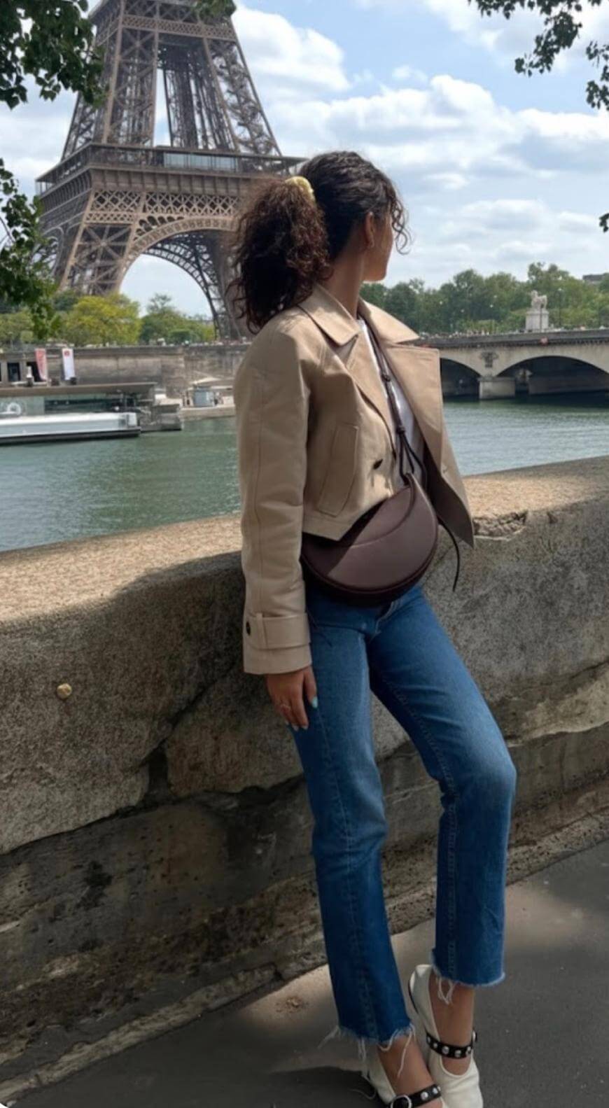 A woman with curly hair wearing a beige jacket and jeans stands by a river, looking toward the Eiffel Tower in Paris on a partly cloudy day.