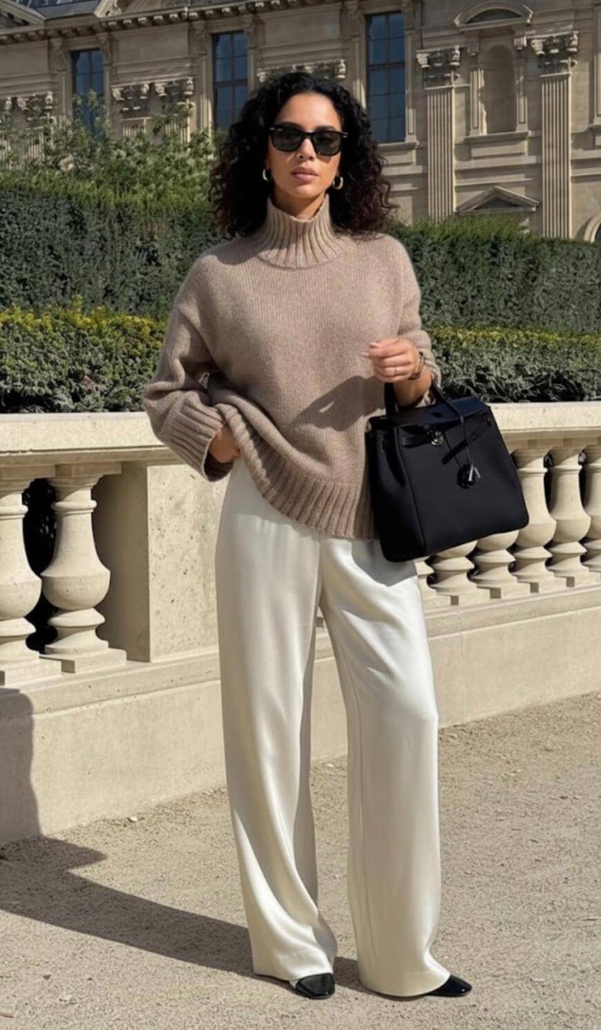 A woman wearing sunglasses, a beige turtleneck sweater, and white wide-leg pants stands outdoors, holding a black handbag. She is in front of a stone railing and a historic building with ornate architecture.
