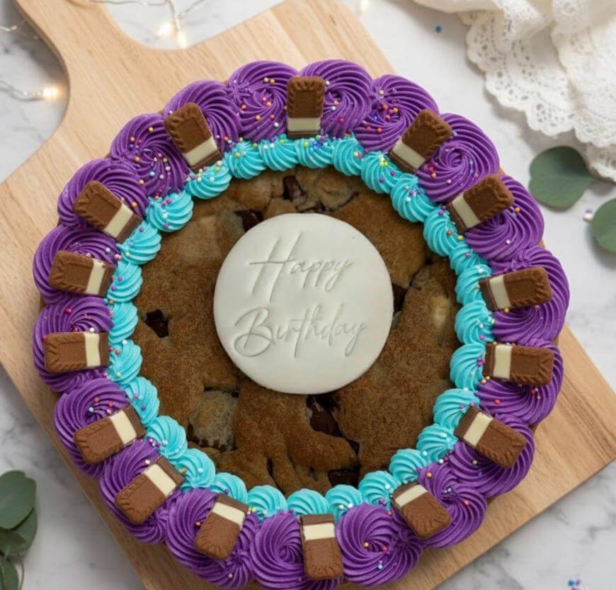 A large cookie cake decorated with purple and blue frosting, mini chocolate bars, and a white fondant disc in the center that says Happy Birthday, displayed on a wooden board.
