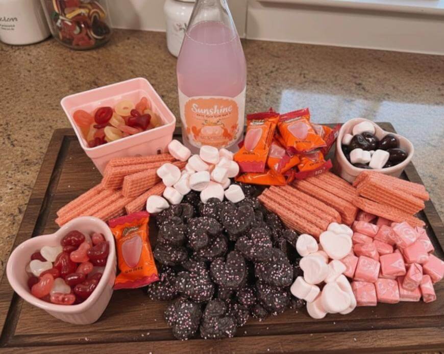 A wooden tray topped with heart-shaped gummies, pink wafers, wrapped candies, pink starbursts, and a pink drink bottle. The candies are arranged in bowls and piles, creating a festive, sweet display.