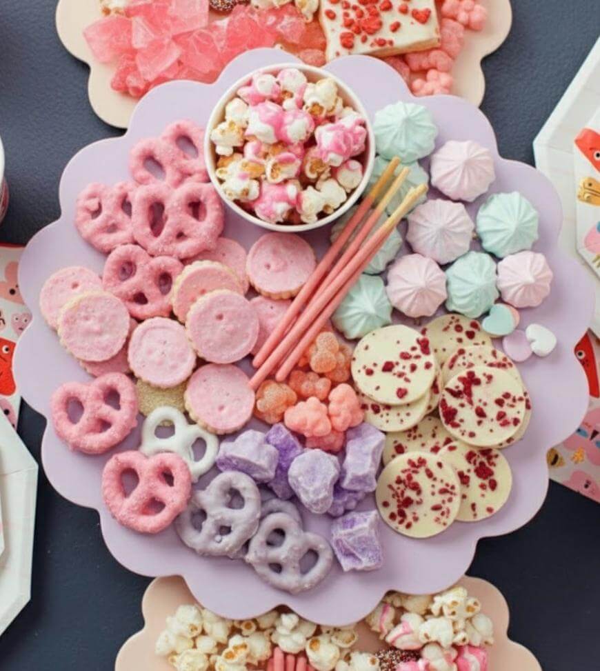 A pastel-colored dessert platter features pink and white chocolate-covered pretzels, heart-shaped cookies, pastel meringues, candy popcorn, wafer sticks, candies, and white chocolate disks with berry pieces on a scalloped tray.