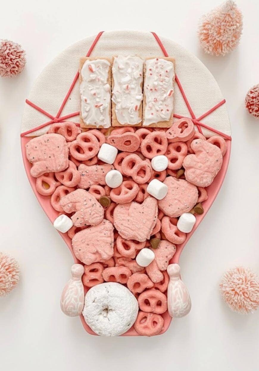 A dessert platter arranged in the shape of a hot air balloon, featuring pink frosted pretzels, frosted animal crackers, mini marshmallows, a powdered donut, and rectangular frosted pastries on top. Pom-poms surround the tray.