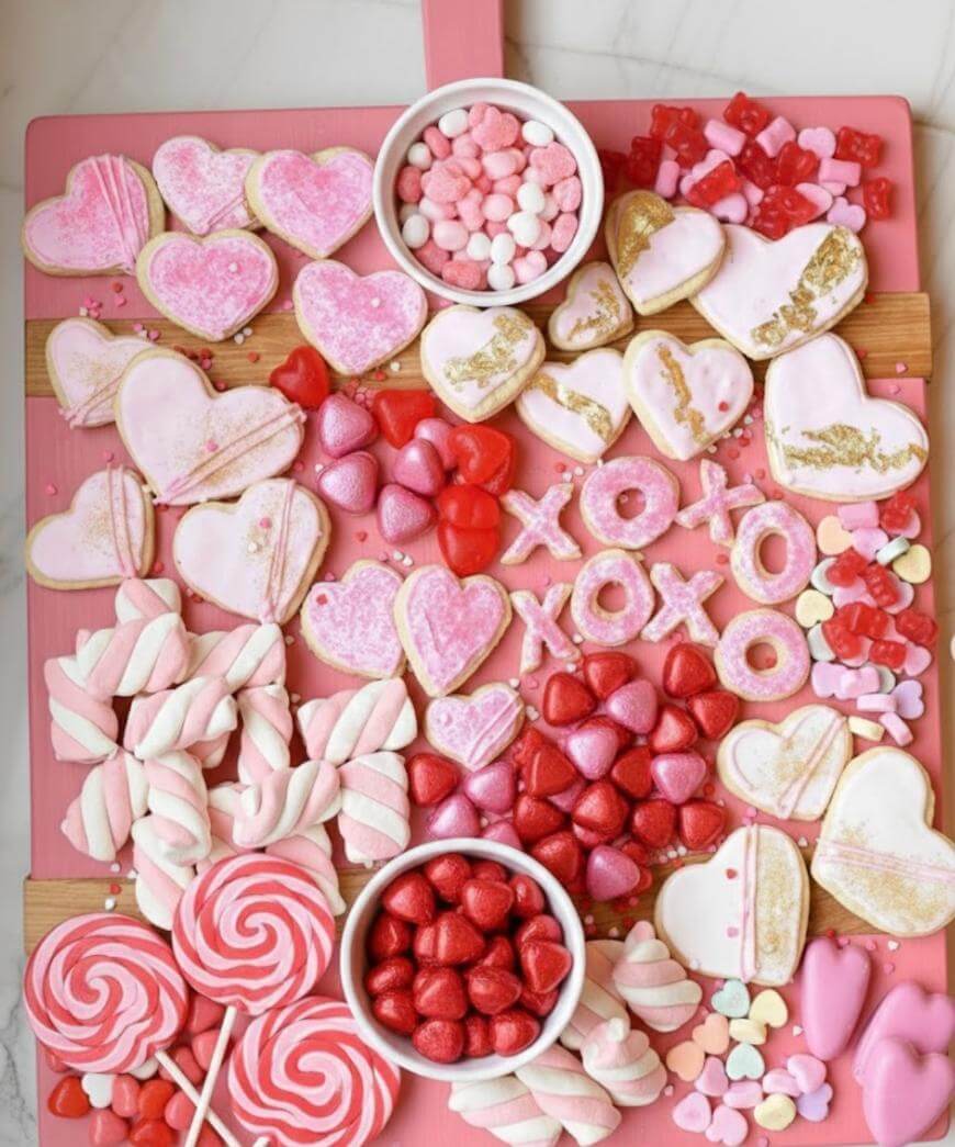 A pink board topped with heart-shaped and XOXO cookies, pink and red candies, marshmallows, lollipops, and bowls filled with small sweets, arranged for a festive Valentines Day treat display.