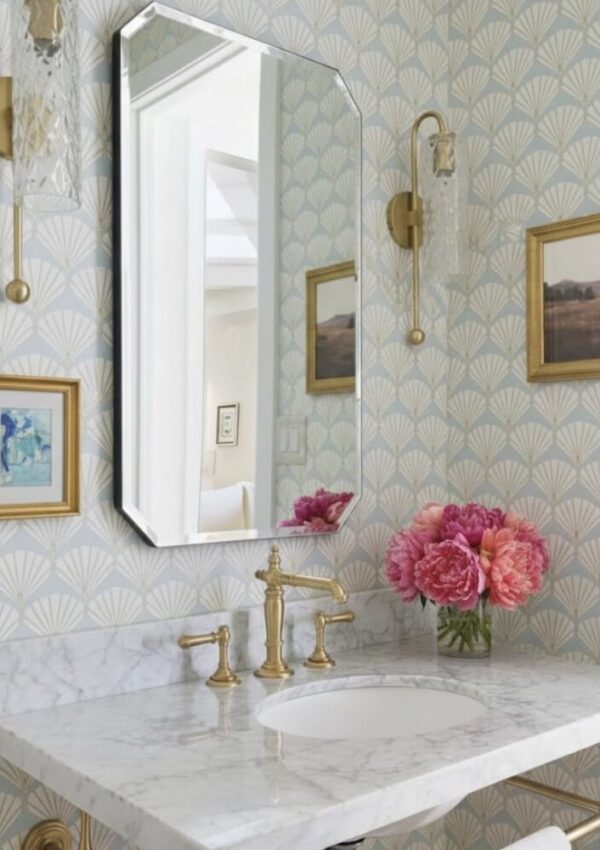 A bathroom with patterned wallpaper, a marble countertop, brass faucet, gold-framed artwork, a geometric mirror, wall sconces, and a vase of pink flowers beside the sink.