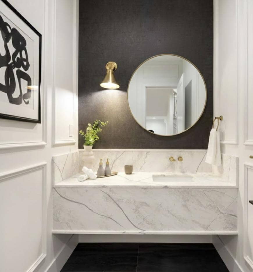 Modern bathroom with a marble sink, round mirror, gold wall sconce, and minimalist decor including a small vase with greenery. Abstract artwork hangs on the white paneled wall to the left.