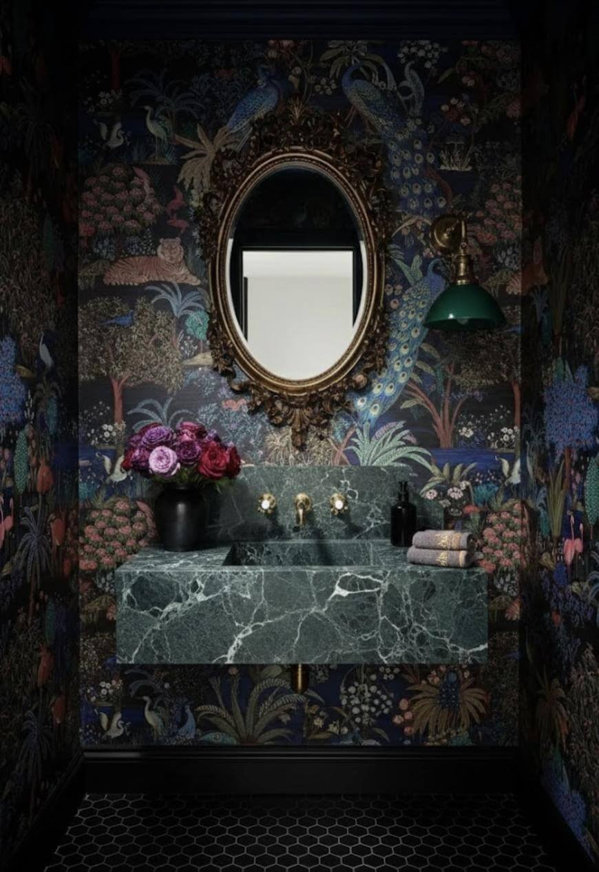A luxurious bathroom features dark floral wallpaper, a marble green sink, a gold-framed oval mirror, a green wall sconce, a vase with purple and red flowers, and neatly folded towels on a black hexagon tile floor.
