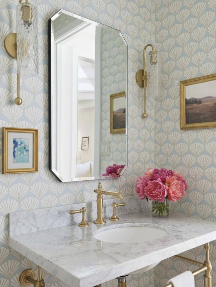 A bathroom vanity with a marble countertop, brass faucet, and an octagonal mirror. The walls have a scallop-patterned wallpaper, gold-framed art, two sconces, and a vase of pink flowers on the counter.