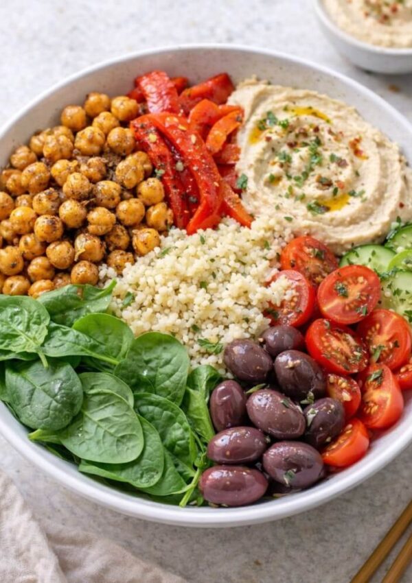 A bowl filled with spinach, roasted chickpeas, red bell peppers, hummus, bulgur, cherry tomatoes, cucumber slices, and Kalamata olives, arranged in colorful sections.