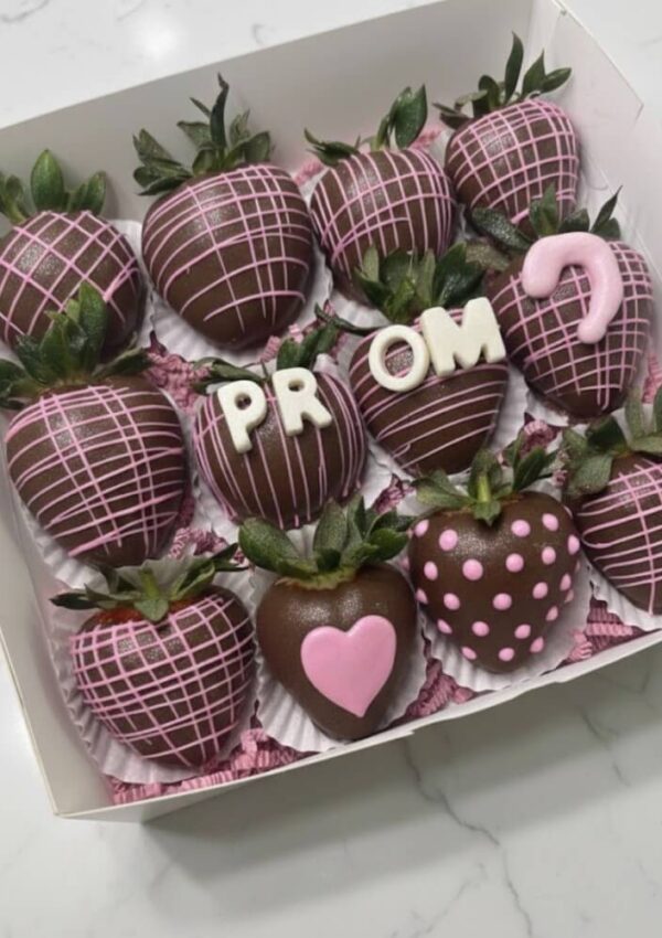 A box of chocolate-covered strawberries decorated with pink icing. Some strawberries spell out PROM? with white and pink letters, while others have heart and polka dot designs.