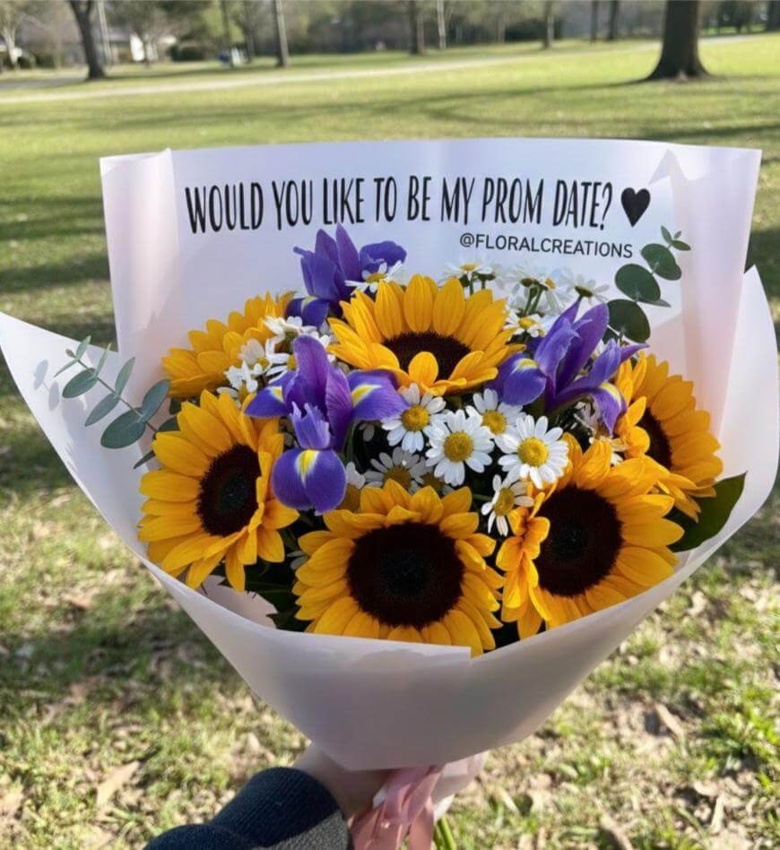 A bouquet of sunflowers, purple irises, and white daisies wrapped in white paper with a note that reads, Would you like to be my prom date? ♥ @floralcreations, held outdoors in a park.
