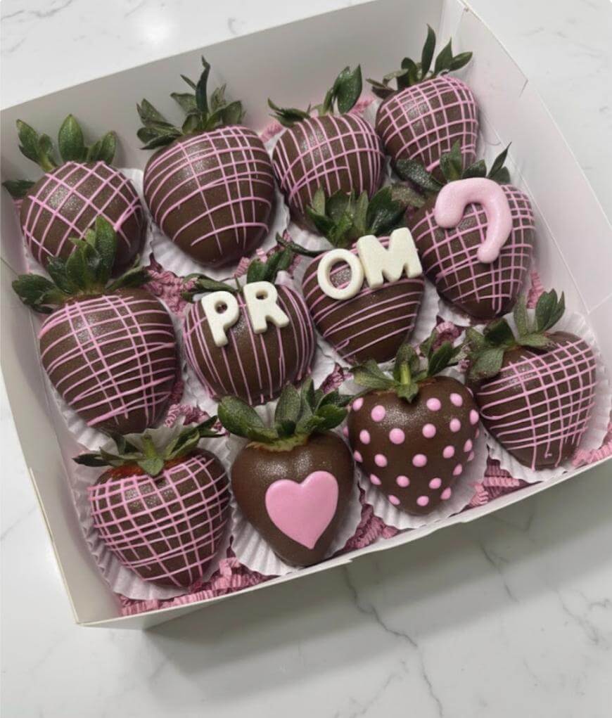 A box of chocolate-covered strawberries decorated with pink icing. Four strawberries spell PROM? with white letters, and others have heart and polka-dot designs. The box sits on a white surface.